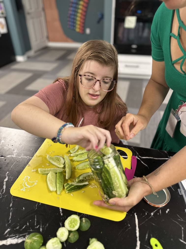 An Onward Adult Day Center participant during a cooking class. She is cutting up pickles and putting them in a jar, with the help of a staff member.